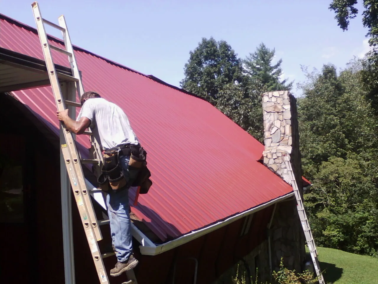 Roofer on a ladder beside a red metal roof and stone chimney — Soffit & Fascia Repair by Roofing Brothers in Sandy Lake